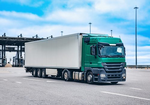 A Green Freight Truck Approaches a Distribution Center with Long Clouds and Overcast Sky in the Background, Highlighting Logistics and Transport Operations