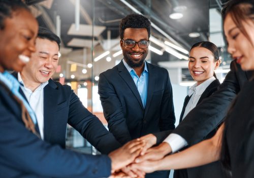 A team of successful businessmen of different nationalities in suits celebrating victory, success while standing in a modern office with their hands clasped together.